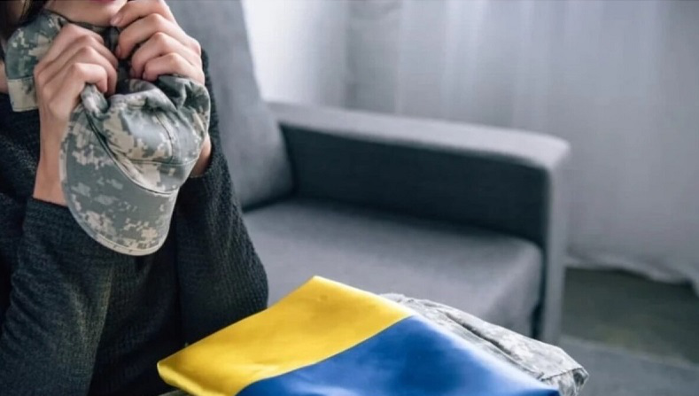 фото жінки, яка сидить на дивані перед прапором України та тримає в руках кепку військового