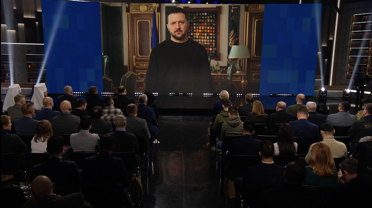 люди в залі сидять перед екраном, на якому транслюється виступ Президента України Володимира Зеленського 