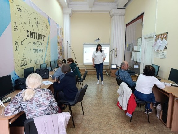 Люди сидять в аудиторії з комп’ютерами під час виступу спікерки, яка стоїть біля проєктора