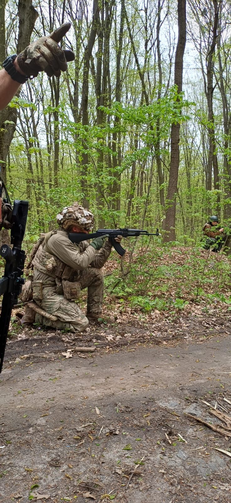 Людина в однострої, присівши, прицілюється, тримаючи в руках зброю