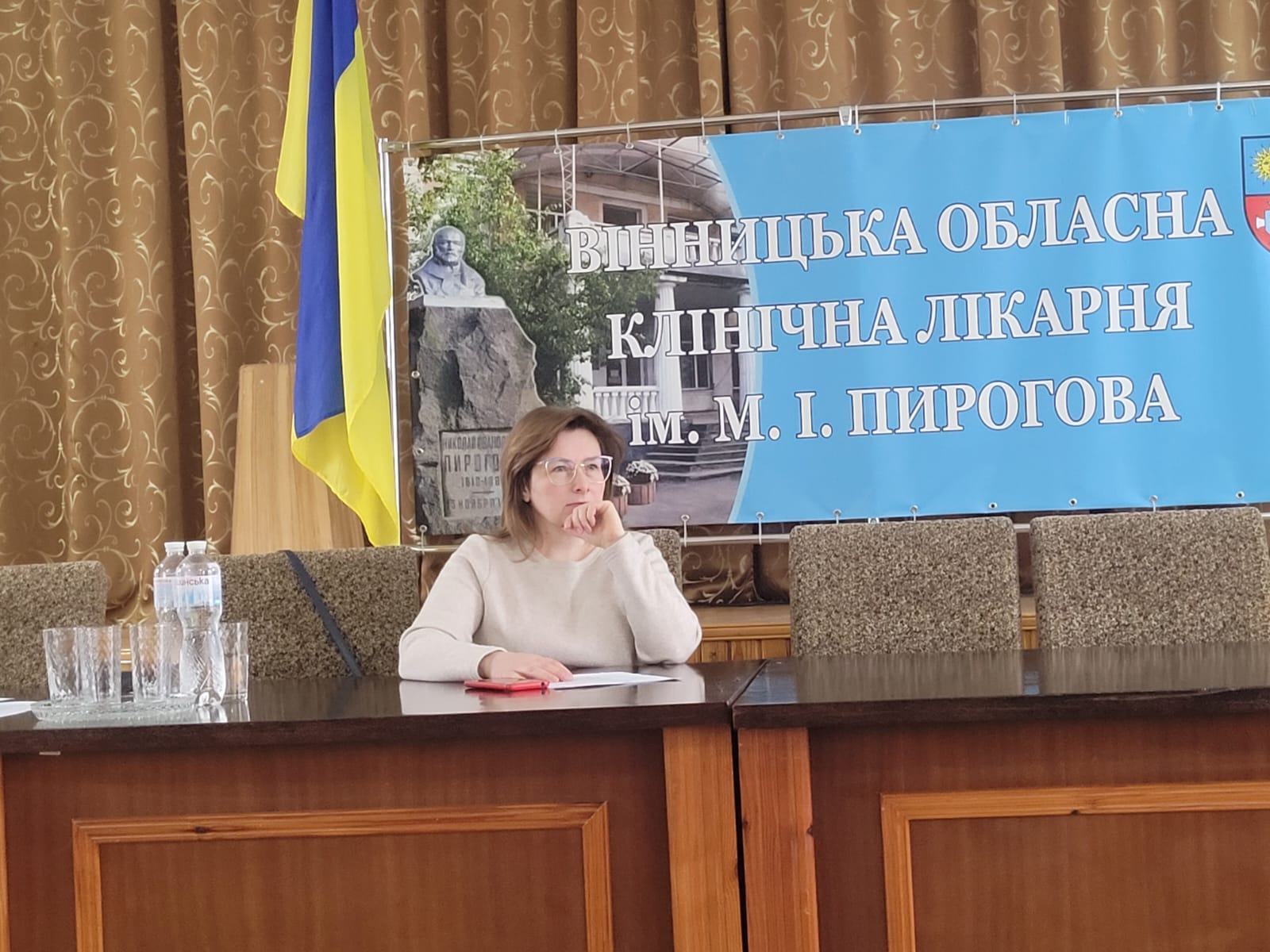 Жінка сидить за столом, за спиною плакат.