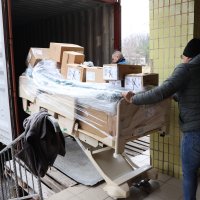 Чоловіки вивозять з вантажного автомобіля медичне ліжко з коробками гуманітарного вантажу