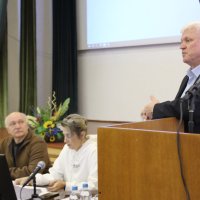 Чоловік за трибуною. Двоє людей сидять у президії