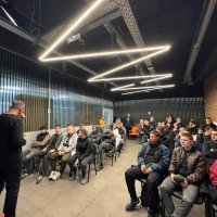 Аудиторія заповнена слухачами, які сидять у рядах. У центрі уваги — доповідач, який виступає перед цією групою.