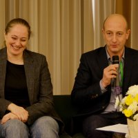 Двоє людей сидят на стільцях, один говорить у мікрофон, пред ними столик на якому стоять квіти