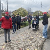 Група з 5–7 осіб, включаючи учасника на візку та людину на милицях, повільно рухається тротуаром, слухаючи гіда.